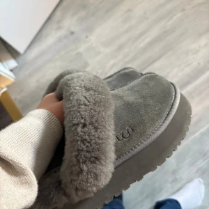 Grå UGG tofflor i fårskinn - Mysiga grå UGG tofflor med fluffig päls runt öppningen och ovandel i mjuk mocka. Perfekta för kalla dagar hemma, med klassisk UGG-logga på sidan och insidan. Sulan är robust och ger bra grepp på golvet.