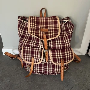Rutig ryggsäck från Daniel Ray - Snygg rutig ryggsäck i vinrött och beige från Daniel Ray. Väskan har läderdetaljer, spännen och snörning upptill. Rymlig insida och två stora fickor framtill. Perfekt för dig som vill sticka ut med en unik och retroinspirerad look.