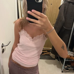 Ljusrosa ribbad linnetopp med spets - Supersöt ljusrosa linnetopp med tunna axelband och ribbad struktur. Toppen har en vacker spetskant längs urringningen och ryggen som ger en romantisk vibe. Perfekt att styla med jeans eller kjol för en chill look.