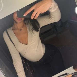 Beige omlottopp med djup v-ringning - Säljer en beige ribbad omlottopp med lång ärm och djup v-ringning. Toppen är figurnära och har en snygg omlottdesign som framhäver midjan. Perfekt att styla med både jeans och kostymbyxor för en trendig look.