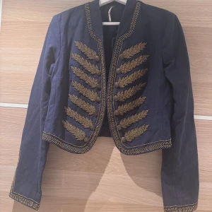 Blå militärinspirerad jacka Free People - Snygg mörkblå militärinspirerad jacka från Free People med guldiga broderier och dekorativa knappar framtill. Jackan är kortare i modellen, har lång ärm och coola detaljer längs kanter och ärmslut. Perfekt för dig som vill sticka ut med en unik look.
