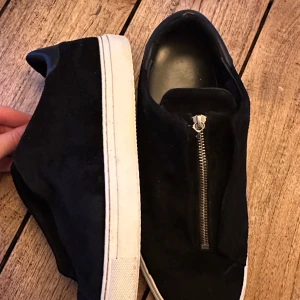 Svarta arigatos i mocka med dragkedja  - Säljer ett par svarta sneakers från Arigato i mjuk mocka med vit platt sula. Skorna har en fast dragkedja. Storlek 44 men skulle rekommendera att du har något större i storlek såsom 45
