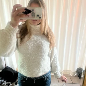 Vit stickad tjock polotröja - Mysig vit stickad polotröja med breda ribbade muddar och hög krage. Tröjan har en loose passform och är lite croppad, perfekt till jeans. Materialet är mjukt och fluffigt, vilket gör den extra skön under kyliga dagar.