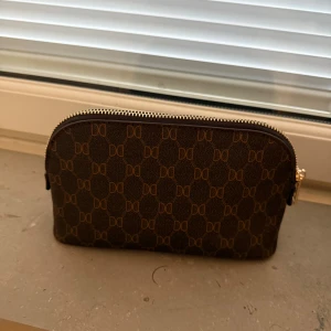  Necessär / Sminkväska - Snygg brun kuvertväska från DonDonna med monogram över hela väskan. Väskan har en smidig dragkedja i guld och är tillverkad i ett tåligt syntetmaterial. Perfekt för att bära det viktigaste med stil.