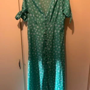 Klänning strl M  - Grön och turkos blommig klänning från Wednesdays’s Girl i strl M. Midi längd, knappar framtill.  Endast använd vid ett tillfälle så jättefint skick. Enda är att tråden har gått upp lite baktill, se sista bilden. Material: polyester  Köpt från Asos. 