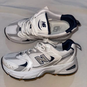 New Balance 530 vita sneakers - Snygga New Balance 530 sneakers i vitt och silver med marinblå detaljer. Skorna har mesh och syntetmaterial, klassisk snörning och en chunky sula med ABZORB-dämpning. Perfekta för dig som gillar sportig och trendig stil.