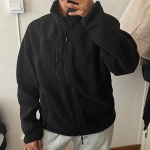 Svart fleece teddyjacka med dragkedja - Mysig svart teddyjacka i fleece med hög krage och hel dragkedja framtill. Jackan har tre praktiska fickor med dragkedja och en loose passform som ger en chill vibe. Perfekt för kalla dagar när du vill vara både bekväm och snygg.