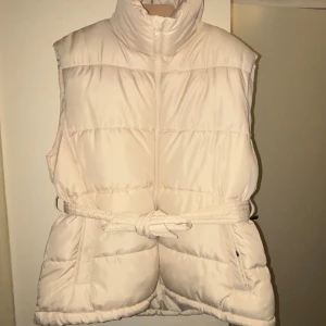 Beige quiltad dunväst med bälte - Snygg beige dunväst med quiltad design och hög krage. Västen har ett knytbälte i midjan som ger en schysst siluett och stängs med dragkedja framtill. Perfekt för lager-på-lager och enkel att styla till olika outfits. Från bikbok helt ny aldrig använt nytt bra skick
