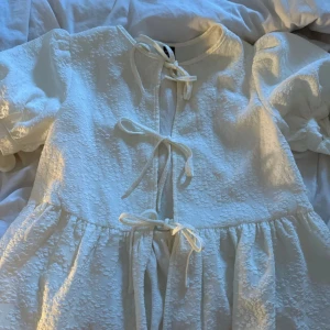 Vit blus med knytband - Superfin vit blus med puffärmar och struktur i tyget. blusen har flera knytband framtill och en lätt volang nedtill. Perfekt för dig som gillar romantiska detaljer och en luftig känsla.