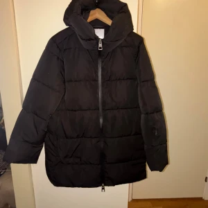 Svart pufferjacka från MNG, M - Svart pufferjacka från MNG i storlek M. Jackan har en stor huva, raka långa ärmar och en kraftig dragkedja framtill. Den är quiltad med breda paneler och har en clean, oversized look som är perfekt för kalla dagar.