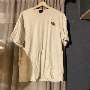 Beige t-shirt med rosbroderi från H&M - Beige t-shirt från H&M i relaxed fit med broderad röd ros på bröstet. Klassisk rund halsringning och korta ärmar. Tillverkad i mjuk bomull, perfekt för en chill och stilren look. Har en fett fläck bredvid rosen och vet ej hur man får bort den