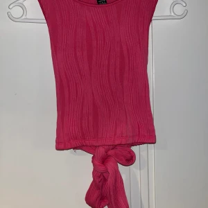 Omlottopp med knyt - Säljer en ribbad rosa topp med omlottdetalj i ryggen och knyt framtill. Toppen är ärmlös och har en tight passform som framhäver figuren. Perfekt för dig som vill ha en trendig och färgstark look.