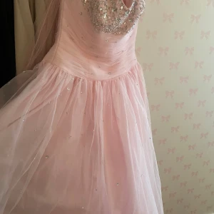 Rosa långklänning med paljetter - Drömmig rosa långklänning med glittriga paljettdetaljer upptill och spridda stenar över tyllkjolen. Klänningen har en romantisk, voluminös kjol och ett vackert draperat liv. Perfekt för dig som vill sticka ut med en sagolik look.