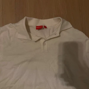 Vit pikétröja från rivyles - Klassisk vit pikétröja från rivyles med korta ärmar och ribbade muddar. Tröjan har en stilren krage och två knappar framtill. Perfekt för en clean och fräsch look, och den röda loggan i nacken ger en snygg detalj.