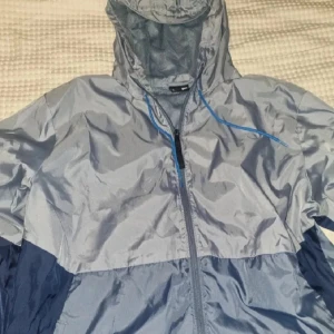 Jacka Under armour  - Vindjacka från Under Armour i olika nyanser av gråblått. Jackan har huva med blå dragsko, hel dragkedja framtill och meshfoder. Inga byten.