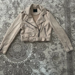 Bershka - Bikerjacka i beige mocka  - Snygg croppad bikerjacka i beige mocka från Bershka. Jackan har klassisk krage, silverfärgade dragkedjor, axelklaffar och bälte i midjan. Perfekt för dig som vill ha en edgy look med stilrena detaljer.
