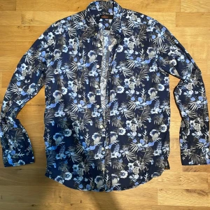 Blommig skjorta från Morris, marinblå - Snygg marinblå skjorta från Morris med blommigt mönster i blått, vitt och grått. Skjortan har klassisk krage, knäppning framtill och långa ärmar. Perfekt för dig som vill sticka ut med en cool och fräsch look. (Storlek L men sitter som M)