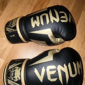 Venum boxningshandskar svart/guld 14oz - Snygga boxningshandskar från Venum i svart och guld med coolt tryck och logga på ovansidan och vid handleden. Handskarna har en robust form och bred kardborrestängning för extra stöd. Perfekta för träning och sparring.