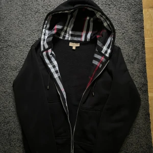 Svart Burberry hoodie - Svart hoodie från Burberry i storlek M med klassiskt rutig foder i huvan i svart, vitt och rött. Tröjan har dragkedja, snörning i huvan och broderad Burberry-logga på bröstet. Perfekt för dig som vill ha en stilren och lyxig streetwear-look.