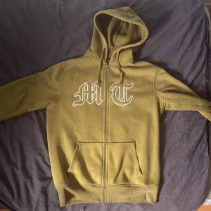 Olivgrön hoodie Active Boys/tuttolente L - Olivgrön hoodie från Active Boys med broderade vita bokstäver 'M T' på bröstet och en stor, diskret örn på ryggen. Tröjan har huva med snörning, dragkedja framtill och två fickor. Insidan är mjuk och fleecefodrad för extra komfort.