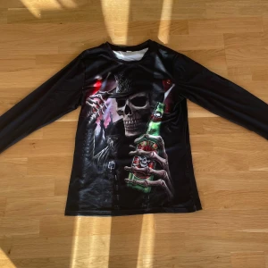 Svart långärmad tröja med dödskalletryck - Svart långärmad tröja med stort tryck av en dödskalle i hatt och läderjacka som håller en grön flaska med texten 'Triple 6'. Röd detalj på ena ärmen. Tröjan har rund hals och är gjord i ett glansigt syntetmaterial. Perfekt för dig som gillar rockig och edgy stil.