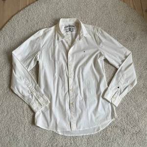 Vit Replay skjorta - Klassisk vit skjorta från Replay Blue Jeans med broderad röd 'R' på bröstet. Skjortan har lång ärm, knappar framtill och en snygg krage. Tillverkad i bomull för en skön och stilren känsla.