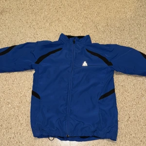 Rahlp louren - Sportig blå vindjacka från Odlo med svarta detaljer och dragkedja framtill. Jackan har hög krage, långa ärmar och justerbar nederkant. Perfekt för utomhusaktiviteter och träning. Tillverkad i lätt och smidigt syntetmaterial.