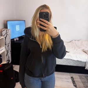 Svart hoodie med dragkedja och huva - Säljer en svart hoodie med dragkedja och stor huva. Tröjan har två fickor framtill och ribbade muddar vid ärmslut och nederkant. Perfekt för chill dagar och enkel att matcha med jeans eller leggings.