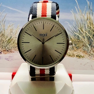 Regal klocka med cool randigt band - Stilren klocka från Regal med stor, rund silverfärgad urtavla och minimalistiska svarta markeringar. Bandet är randigt i marinblått, vitt och rött och tillverkat i slitstark syntet. Perfekt accessoar för dig som gillar såväl  klassisk som sportig still. Säljes i fungerande skick!