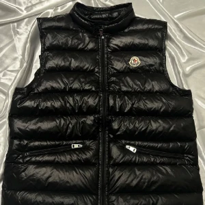 Svart dunväst från Moncler - Svart glansig dunväst från Moncler med hög krage, två dragkedjefickor och klassisk logotyp på bröstet. Västen har quiltad design och stängs med dragkedja framtill. Perfekt för lager-på-lager och streetstyle.