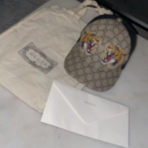 Gucci tiger keps - Säljer en exklusiv Gucci keps med klassiskt GG-monogram i beige och brunt, prydd med två broderade tigrar framtill. Baksidan har justerbart band i grönt och rött. Kepsen är tillverkad i bomull och mesh för skön passform. Perfekt för dig som vill sticka ut med lyxig streetstyle samt dustbag ingår med kvittot och den är i storlek S. Helt ny, har inte använt den alls. Den är helt svår och få tag på då man inte kan köpa den nånstans alls eftersom Gucci har slutat producera denna modellen för längese