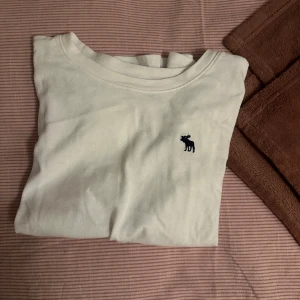 Vit t-shirt från Abercrombie Kids - Vit t-shirt från Abercrombie Kids med klassisk rund hals och korta ärmar. Tröjan har en liten mörkblå älg-logga broderad på bröstet. Tillverkad i mjuk bomull, perfekt för en clean och enkel stil.