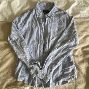 Randig skjorta från Polo Ralph Lauren - Klassisk blå- och vitrandig skjorta från Polo Ralph Lauren med broderad logga på bröstet. Skjortan har lång ärm, knappar framtill och är tillverkad i bomull. Perfekt för en clean och stilren look.