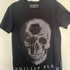 Svart Philipp Plein t-shirt med döskalle - Svart t-shirt från Philipp Plein med stor glittrig döskalle framtill och logga både fram och bak. T-shirten har korta ärmar och är tillverkad i mjuk bomull. Snygg detalj med paljetter som ger extra edge till looken.