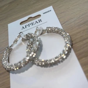 Glittriga hoops från Appear - Säljer ett par stora, glittriga hoops från Appear. Örhängena är silverfärgade och täckta med gnistrande stenar som verkligen fångar ljuset. Perfekta för dig som vill sticka ut och addera extra bling till din look.
