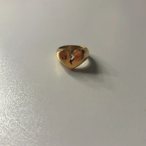 Guldfärgad ring med brustet hjärta - Cool ring 💔 i guldfärg med ett brustet hjärta som motiv. Ringen har en chunky design och är perfekt för dig som vill sticka ut med en unik accessoar. Passar både till vardagslooken och när du vill addera lite edge till din stil.