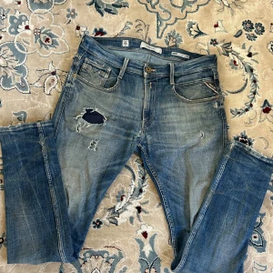 Replay Anbass slitna blå jeans 32 - Säljer ett par Replay Anbass jeans i blå tvätt med slitningar och hål på låret för en cool, avslappnad look. Klassisk femficksmodell med dragkedja och Replay-logga på fickan. Slim fit och bekvämt material i bomull. Perfekta för dig som gillar streetstyle.