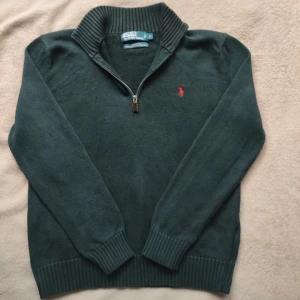 Mörkgrön half zip tröja från Polo Ralph Lauren - Mörkgrön stickad tröja från Polo Ralph Lauren i 100% bomull. Tröjan har en ribbad hög krage med half zip-dragkedja och den klassiska röda broderade loggan på bröstet. Långärmad modell med ribbade muddar och nederkant. Perfekt för lager-på-lager-stil.