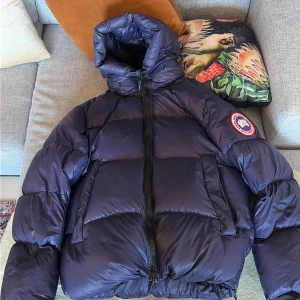Mörkblå pufferjacka från Canada Goose - Säljer en mörkblå pufferjacka från Canada Goose med hög krage och stor huva. Jackan har en tydlig logotyp på ärmen, dragkedja framtill och stora, vadderade partier för extra värme. Perfekt för kalla vinterdagar och riktigt snygg streetstil. Pris kan diskuteras