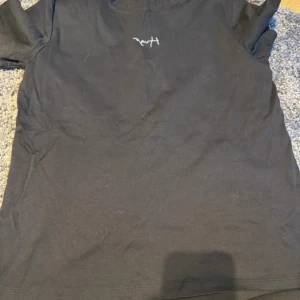 Svart t-shirt med liten vit text XS - Svart t-shirt i storlek XS med en liten vit text på bröstet. Klassisk passform med korta ärmar och rund halsringning. Perfekt basic-plagg att ha i garderoben. Materialet känns som bomull och är mjukt mot huden.