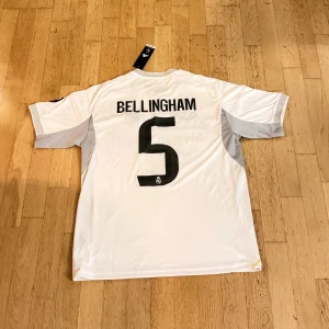 Real Madrid Bellingham tröja Adidas L - Säljer en officiell Real Madrid fotbollströja från Adidas med Bellingham och nummer 5 på ryggen. Tröjan är vit med svarta detaljer, gula inslag och klubbmärke på bröstet. Tillverkad i lätt funktionsmaterial (AEROREADY) och har korta ärmar samt klassiska Adidas-ränder på axlarna.