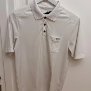 Vit sportig pikétröja från Oakley - Snygg vit pikétröja från Oakley med klassisk krage, tre knappar och liten bröstficka med Oakley-logga. Tillverkad i 100% polyester som ger en lätt och sportig känsla. Perfekt för dig som gillar stilren och enkel design. Storlek M.