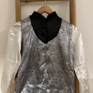 Halloween kläder. Silvrig väst med spindelnätsmönster - Cool silvrig väst med svart krage och detaljer, täckt av spindelnätsmönster och små spindlar. Västen har glansiga vita ärmar och är gjord i ett syntetmaterial som ger en festlig känsla. Perfekt för temakvällar eller halloween.