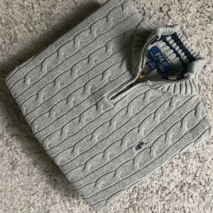 Grå stickad tröja Polo Ralph Lauren - Kabelstickad grå tröja från Polo Ralph Lauren med halv dragkedja och broderad logga på bröstet. Tröjan har ribbade muddar och krage med marinblå rand. Perfekt för lager-på-lager och snygg till jeans.