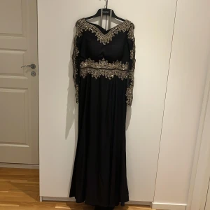 Svart långklänning med guldspets - Elegant svart långklänning med vackra guldiga spetsdetaljer längs halsringning, midja och ärmar. Klänningen har långärmade spetsärmar och en klassisk, flytande siluett. Perfekt för bal eller festliga tillfällen när du vill sticka ut.