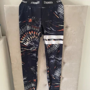 Mönstrade leggings från Stronger - Snygga svarta leggings från Stronger med färgglatt abstrakt mönster i vitt, orange och grönt. De har bred resår i midjan med logga och två vita ränder på ena benet. Perfekta för träning eller chill. Materialet är stretchigt och följsamt. Tror det är storlek XS💞
