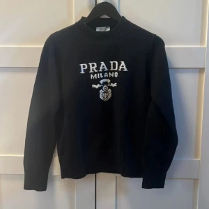 Svart stickad tröja från Prada - Svart stickad tröja från Prada med vit broderad logga och emblem på bröstet. Tröjan har rund halsringning med bred ribbad kant och långa ärmar. Perfekt för dig som vill ha en klassisk och lyxig look med streetvibe.