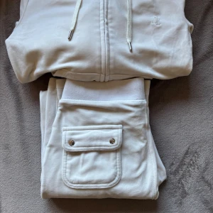 Vita mjukissett från Juicy Couture - Vit mjukisett från Juicy Couture i mjuk velour med bred resår i midjan och en stor ficka med lock och silverfärgade knappar bak. Klassisk broderad JC-logga och relaxed fit. Perfekta för chill dagar eller när du vill vara extra comfy.