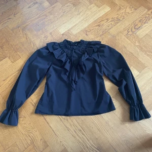 Svart volangblus från Gina Tricot - Supersnygg svart blus från Gina Tricot med volangdetaljer längs bröstet och axlarna. Hög krage med rynk och knytning fram, samt långa ärmar med volang vid ärmslut. Perfekt för dig som gillar en trendig och romantisk look.