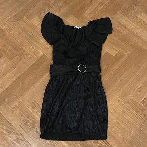 Svart kort klänning med volang  - Supersnygg svart kort klänning med markerad midja och bälte med glittrande spänne. Klänningen har volangdetaljer vid axlarna och en v-ringad omlottfront. Perfekt för dig som vill sticka ut med en elegant och trendig look.